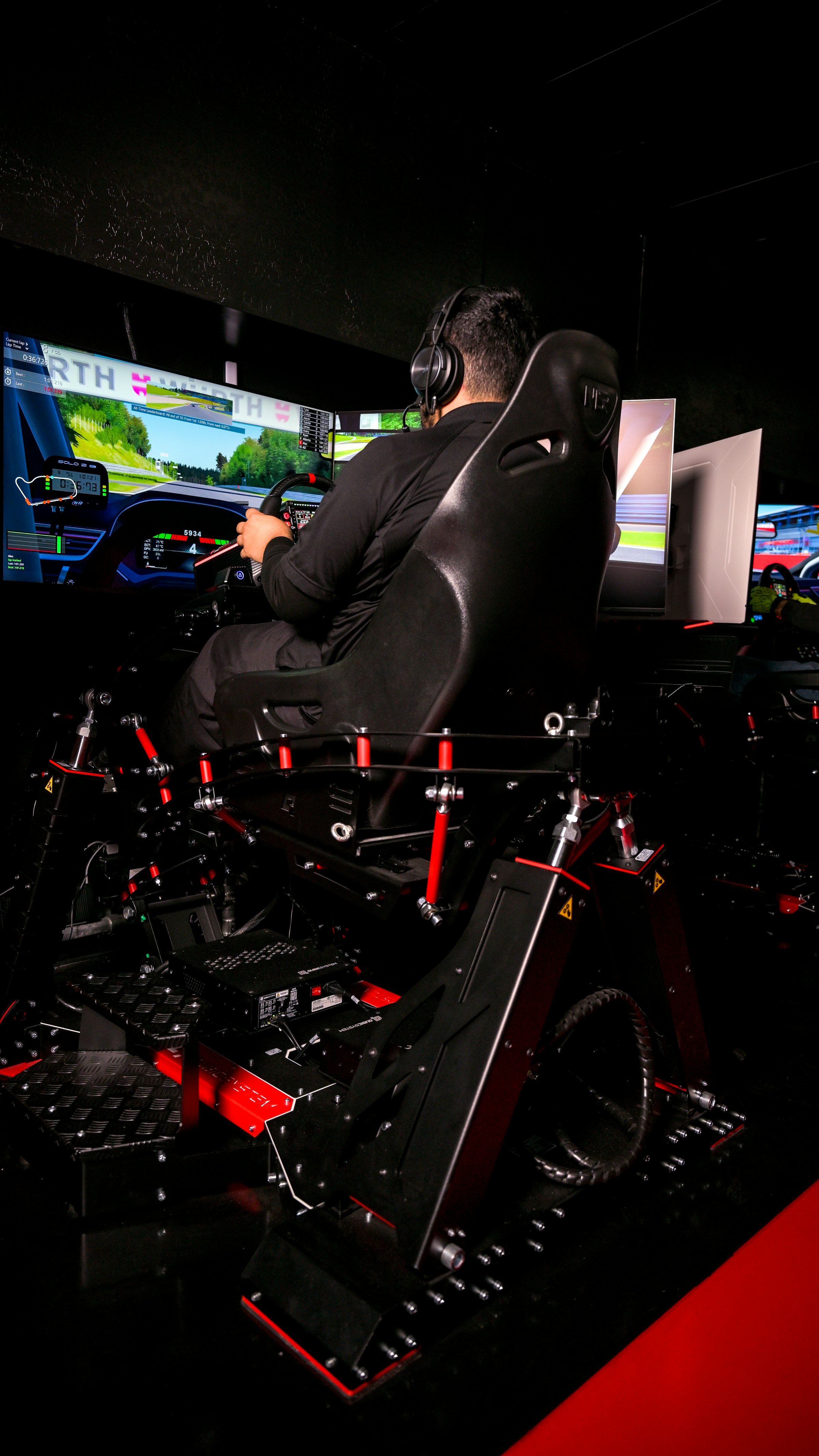 A sim racer in a cockpit rig facing three monitors, gripping a wheel